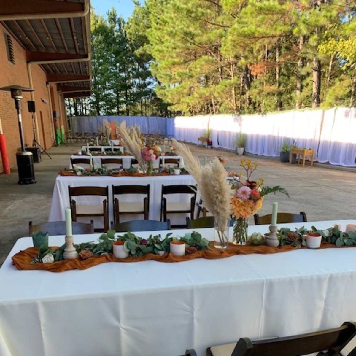 A series of tables are set up outside, decorated with floral arrangements and candles, under a backdrop of trees.