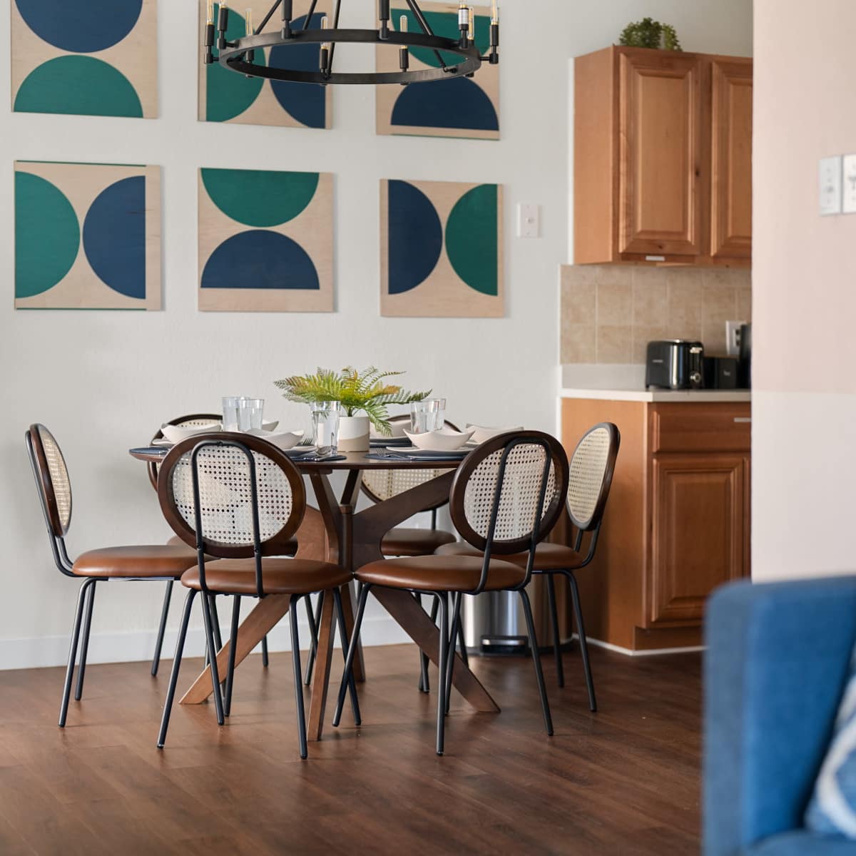 A modern dining area features a round table surrounded by four chairs, with geometric artwork on the wall and a kitchen visible in the background.