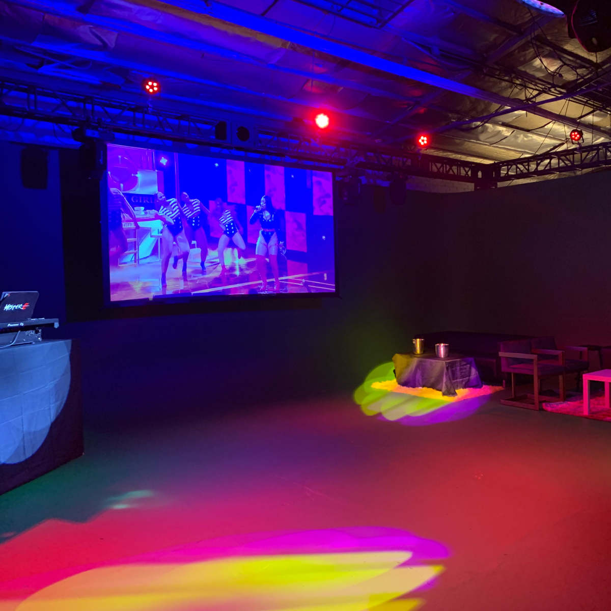A dimly lit event space features a large screen displaying dancers, with colorful lights illuminating the floor and furniture.