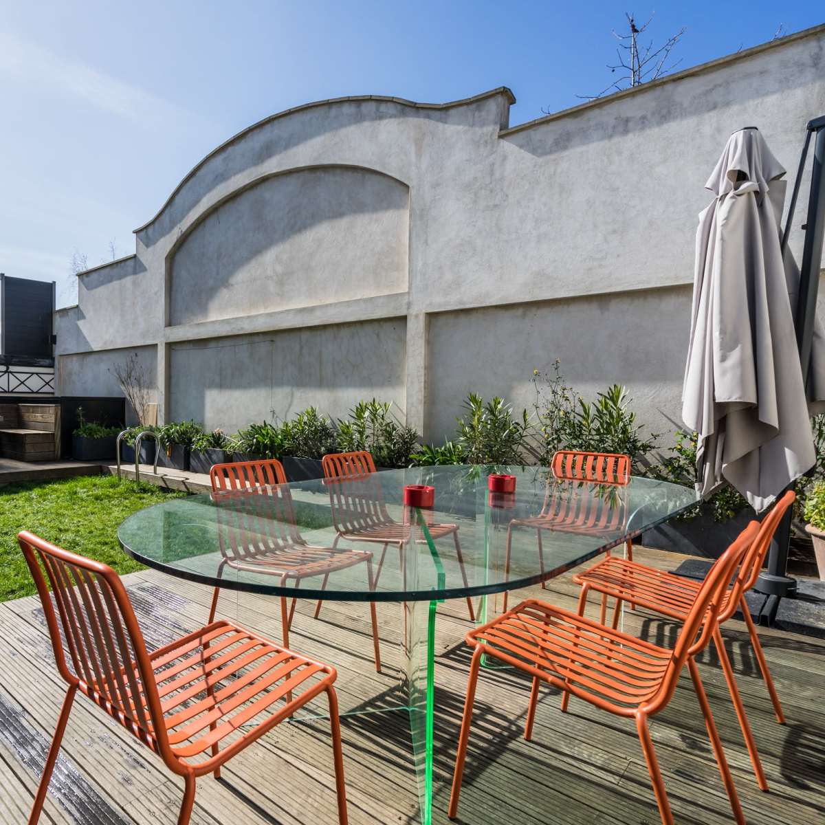 A rooftop terrace features a glass table surrounded by orange chairs, with a green lawn and seating area against a backdrop of a wall.