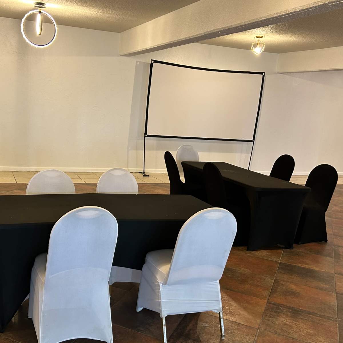 A spacious room features several tables with black and white tablecloths, a large screen at the back, and a few chairs arranged nearby.
