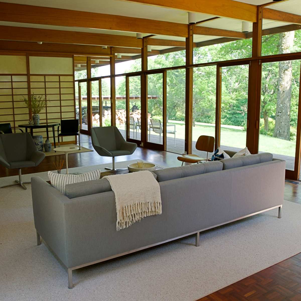 A modern living room features a gray sofa, wooden furniture, and large windows that overlook a green landscape.