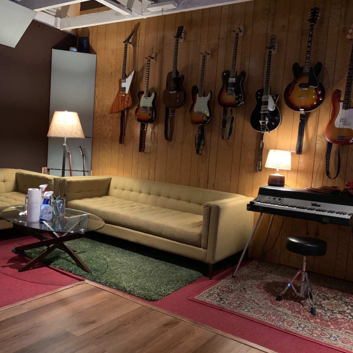 A cozy music studio with several guitars hanging on a wooden wall, a beige sofa, a lamp, a keyboard on a stand, and a rug on the floor.