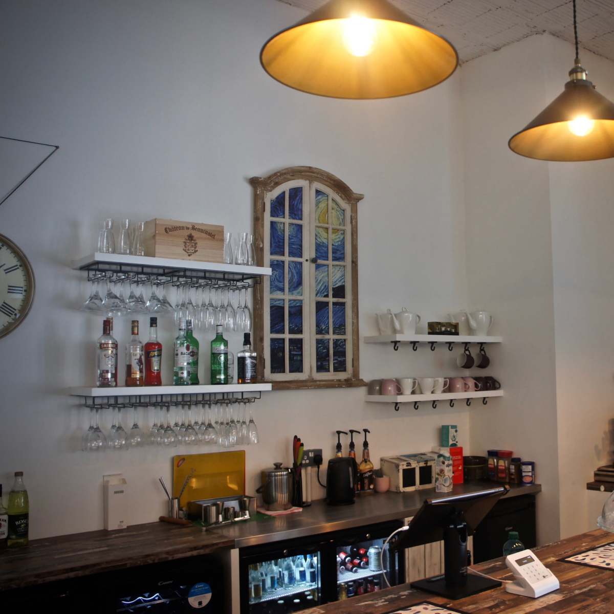 A café interior with a wooden counter, multiple shelves holding glassware and bottles, and pendant lights hanging overhead.