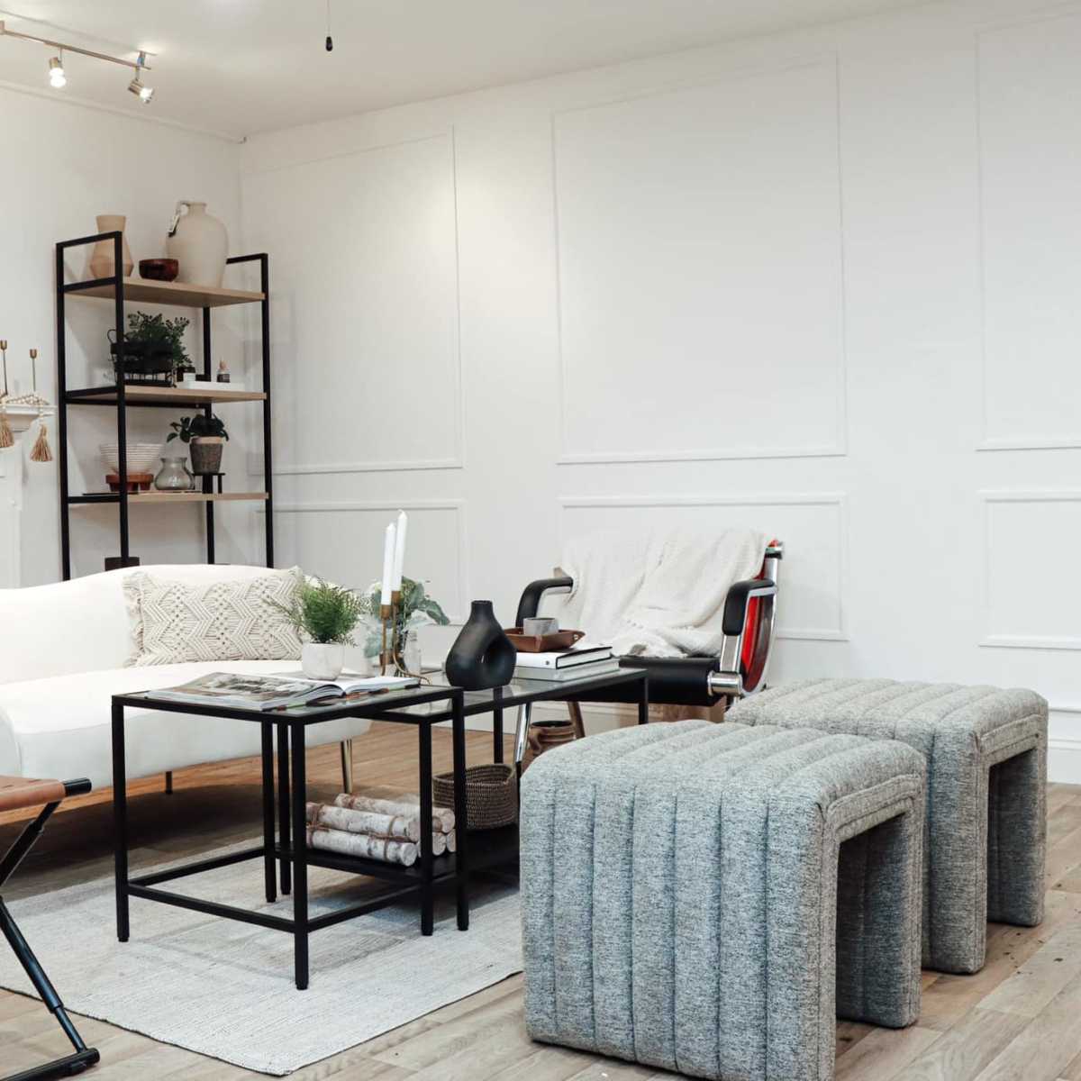A bright living room featuring a white sofa, a black coffee table, and two gray upholstered stools, with shelves and plants in the background.