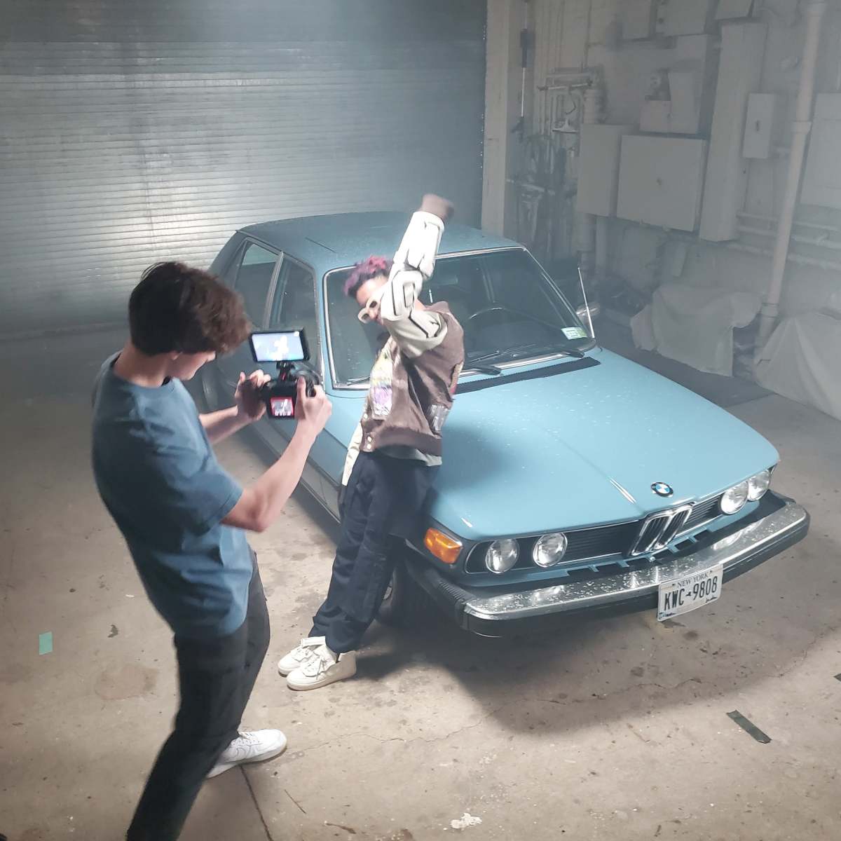 A person films another person posing next to a vintage BMW in a garage filled with smoke.