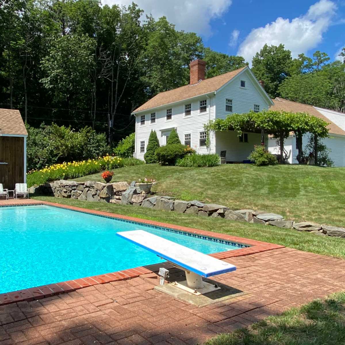 A swimming pool with a diving board is situated in a grassy backyard next to a white two-story house surrounded by trees.