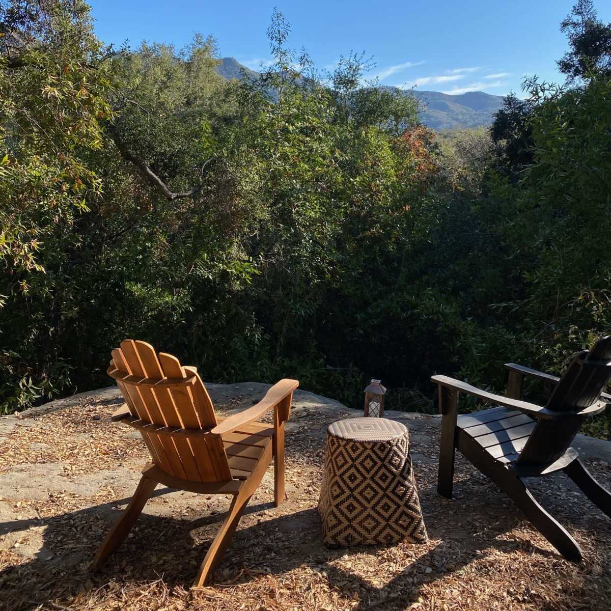 Two chairs sit on a stone patio surrounded by lush greenery, facing a scenic view of distant mountains under a clear blue sky.
