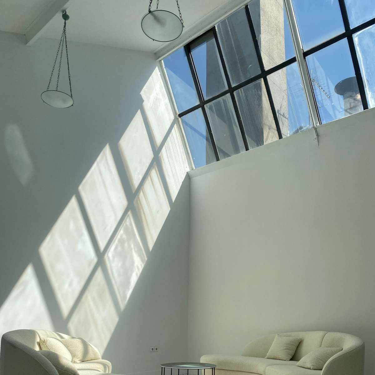 A minimalist living space with white furniture and large windows casting shadows on the tiled floor.