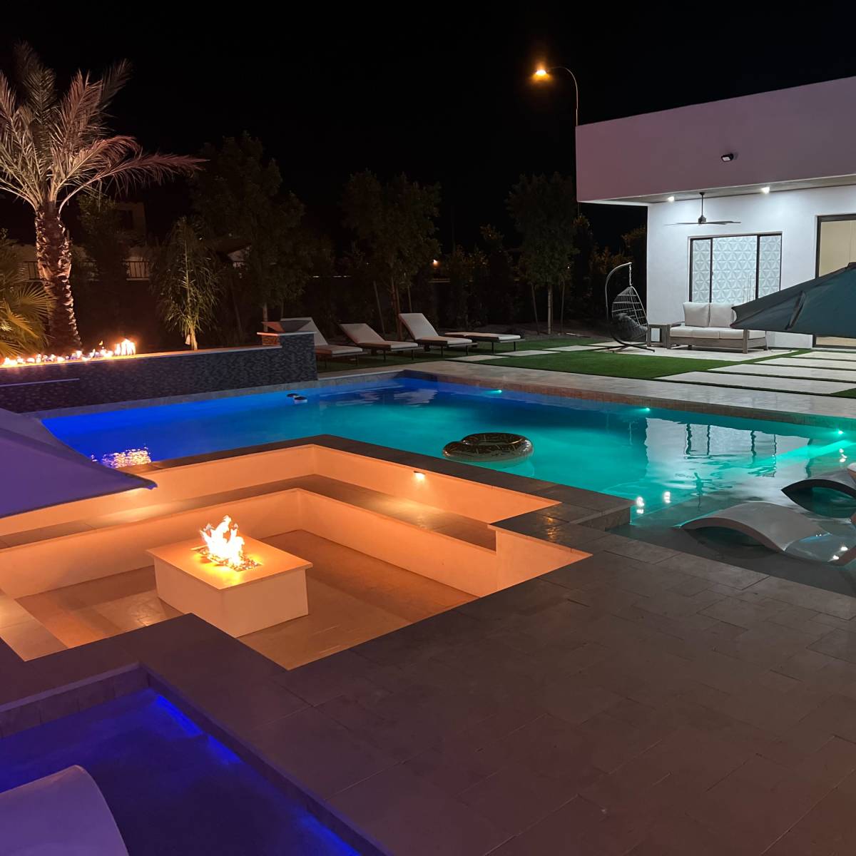 A modern outdoor pool area at night, featuring illuminated water features, lounge chairs, and a fire pit.