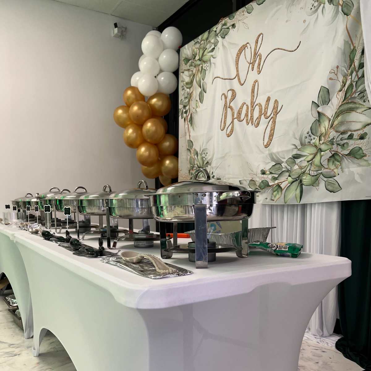 A buffet table is set up with chafing dishes, accompanied by a backdrop featuring the text "Oh Baby" and decorative balloons in gold and white.
