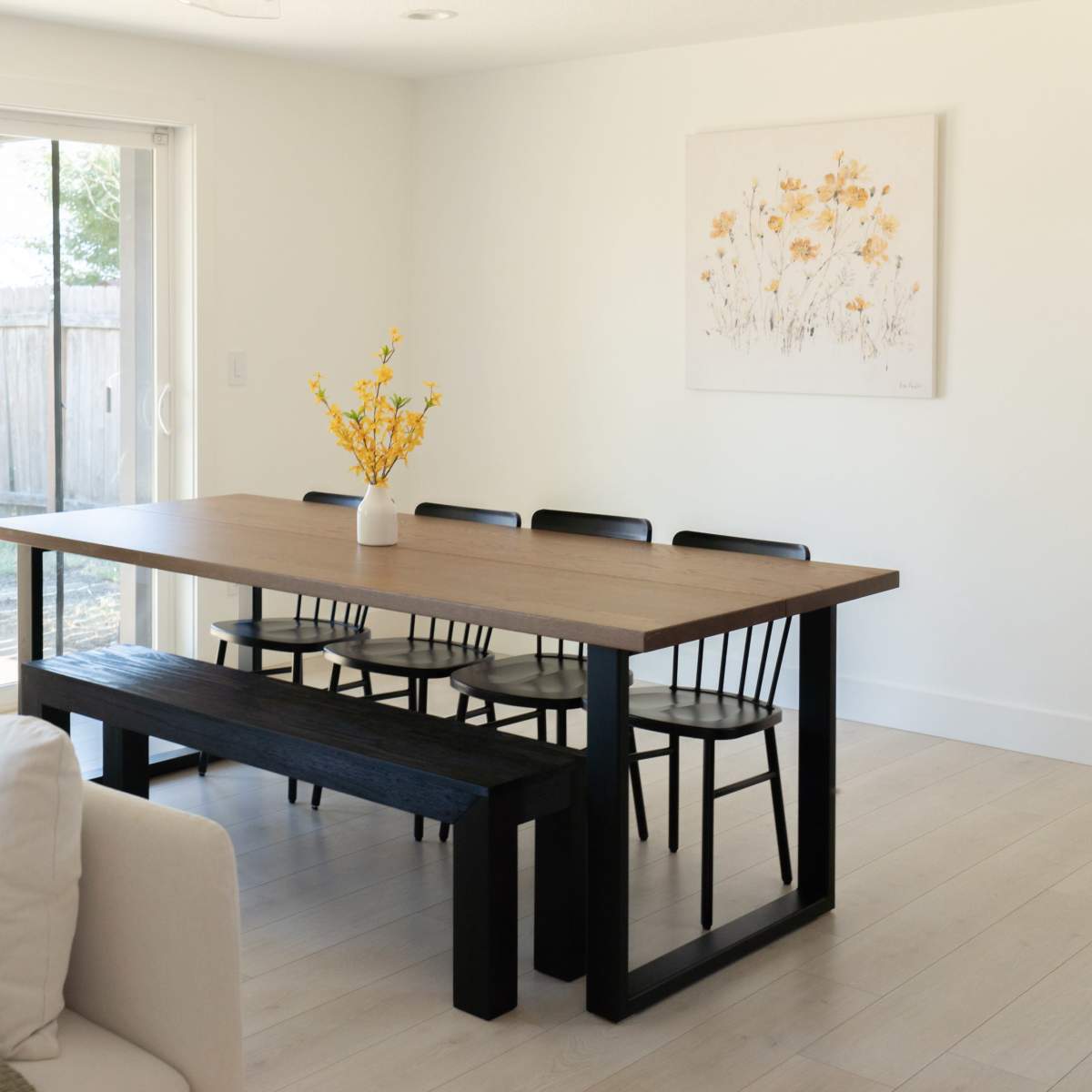 A modern dining area features a wooden table with six black chairs and a bench, complemented by a vase of yellow flowers, against a backdrop of sliding glass doors leading to an outdoor space.