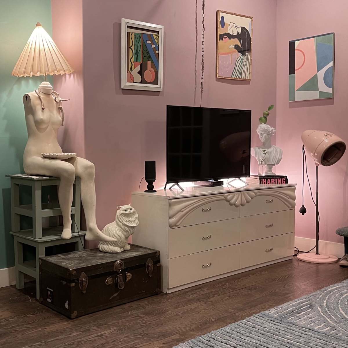 The scene features a cozy room with pastel-colored walls, a television on a white dresser, a mannequin, a decorative lamp, and various framed artworks.