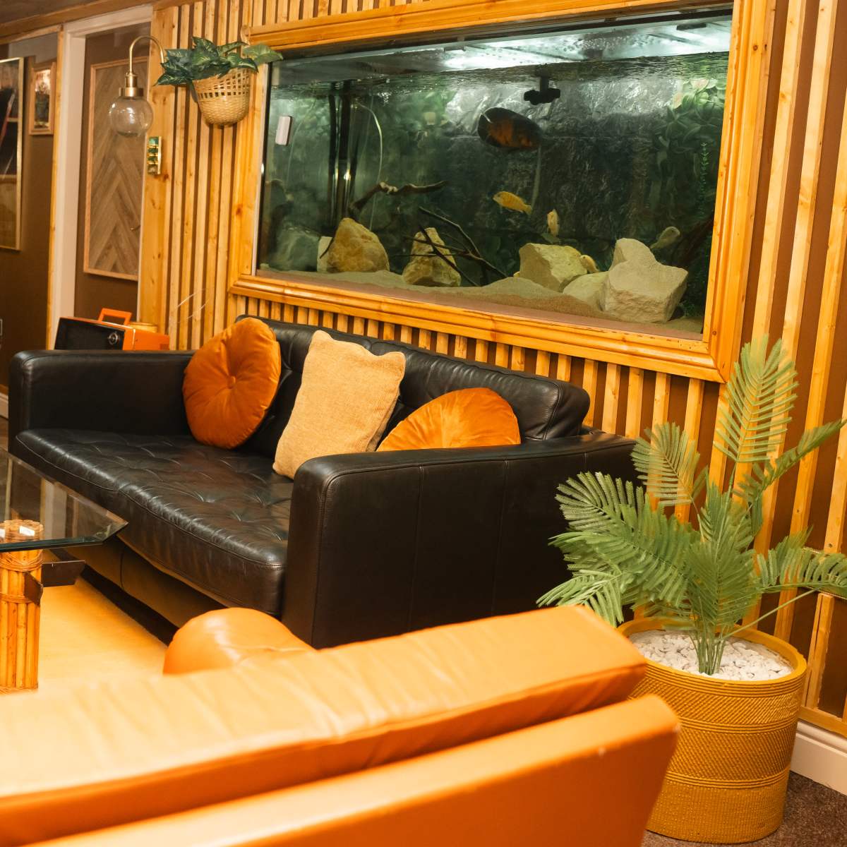 A cozy living room with a black sofa, orange accent cushions, a coffee table, and an aquarium along a wooden-paneled wall.