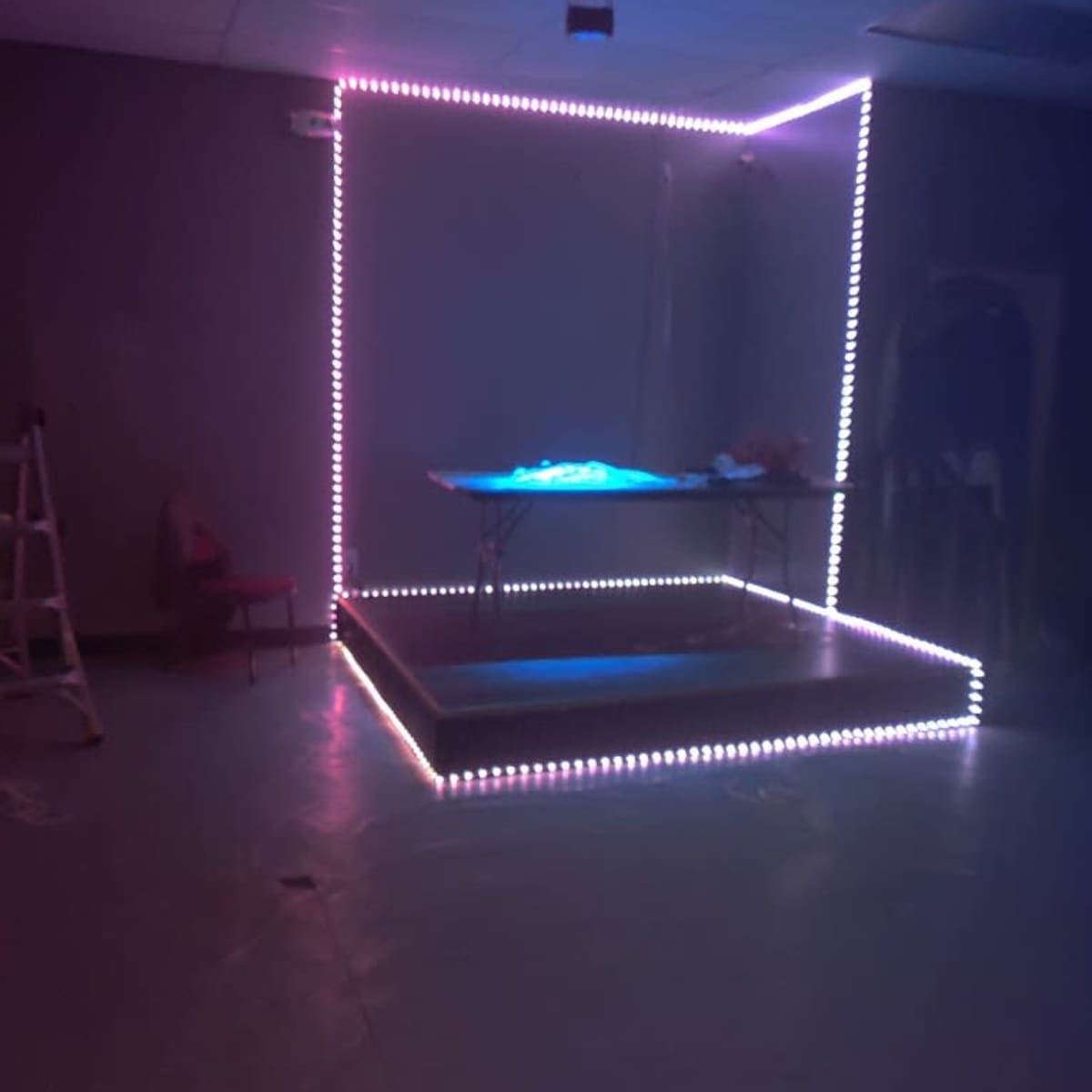 A brightly lit stage area with LED strip lights outlining a rectangular platform and a table in a sparsely furnished room.