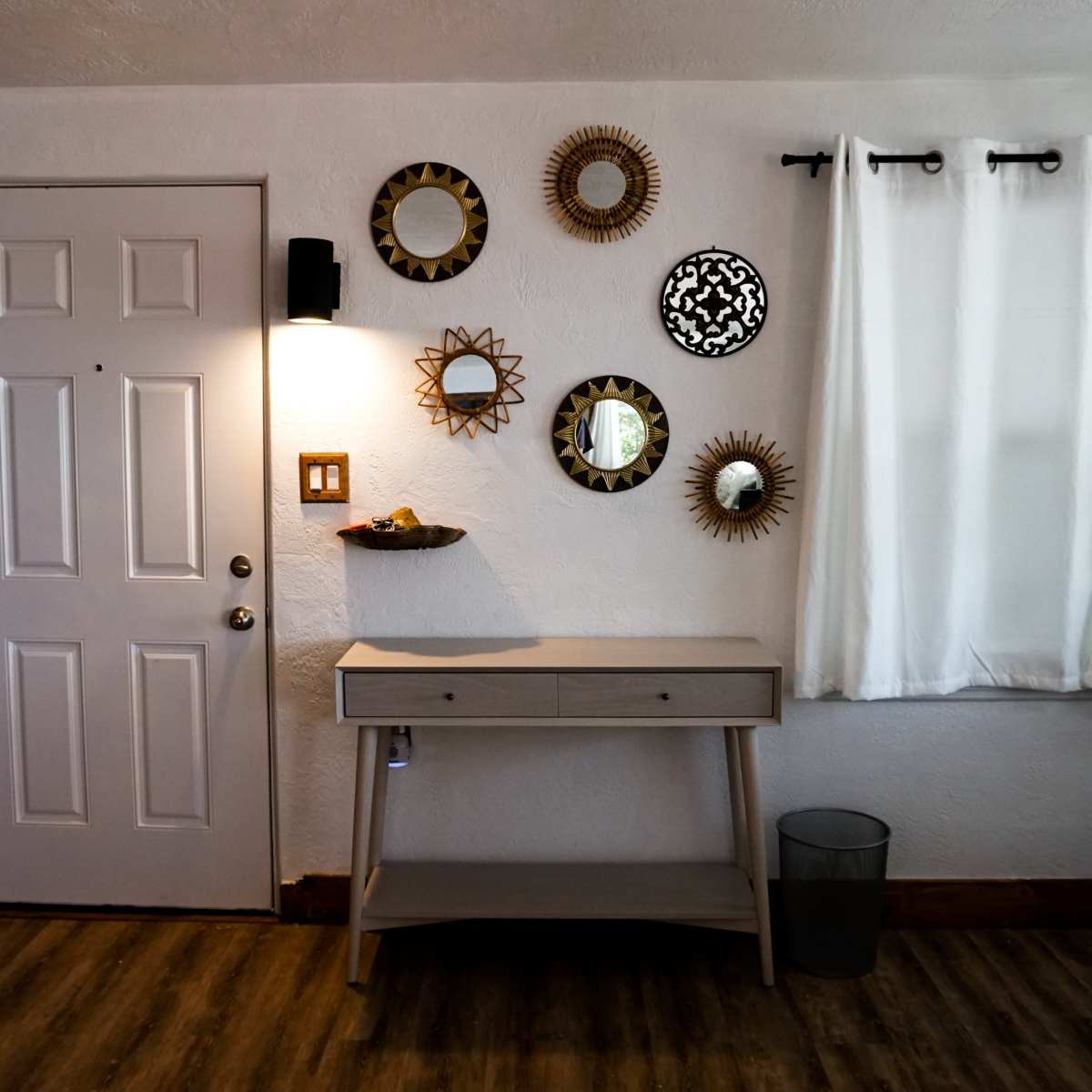 A light-colored wooden table is positioned against a wall featuring decorative mirrors and a door on the left, with a white curtain partially covering a window.