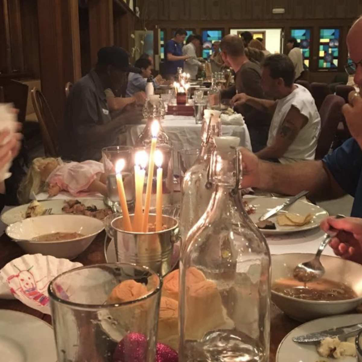 A long dining table is filled with people eating dinner in a warmly lit room, with candles and various food items visible.
