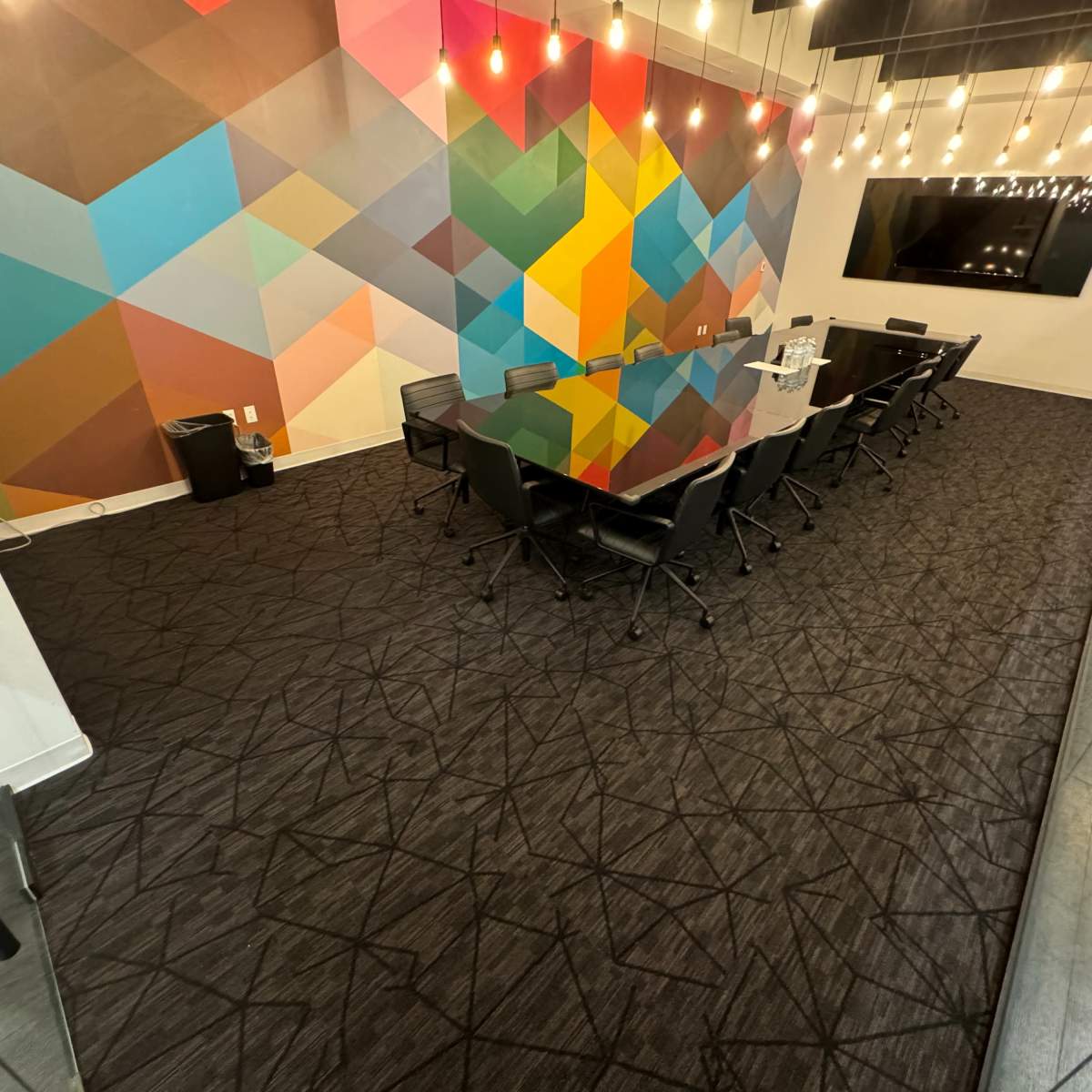 A modern conference room features a large black table surrounded by chairs, with a colorful geometric mural on the wall and decorative lighting above.