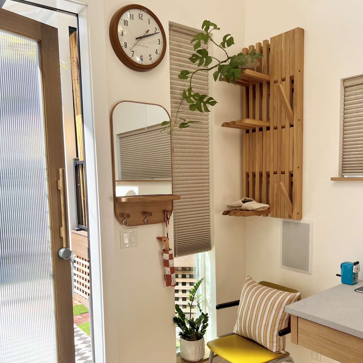 A small, well-organized room features a clock, a mirror, wooden shelves, and a plant, with a door leading outside to a patio area.