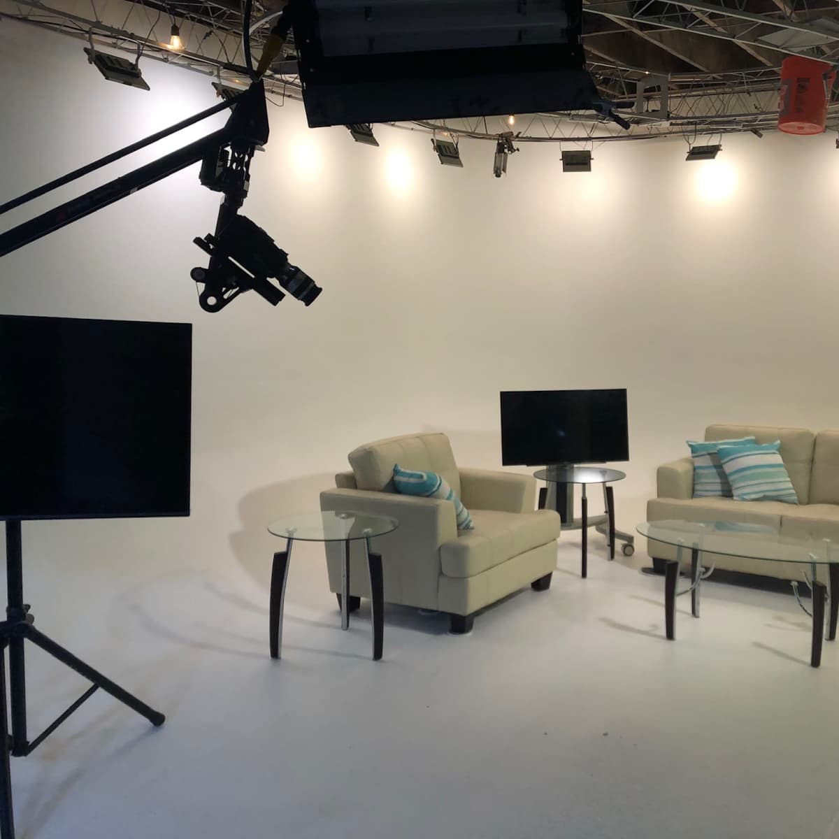 A well-lit studio setting features a beige sofa and armchair with blue and white striped pillows, two glass tables, and a television on a stand.
