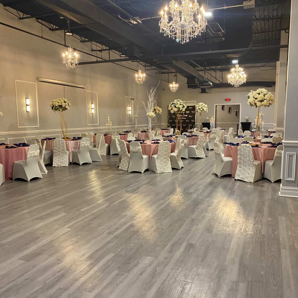 A banquet hall set up with tables covered in pink and white linens, adorned with floral centerpieces and chandeliers hanging from the ceiling.