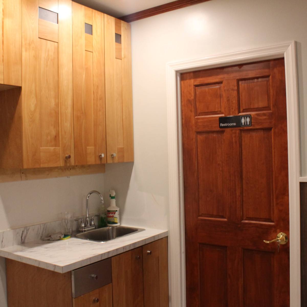A small kitchenette area with wooden cabinetry, a stainless steel sink, and a door labeled "Restroom."