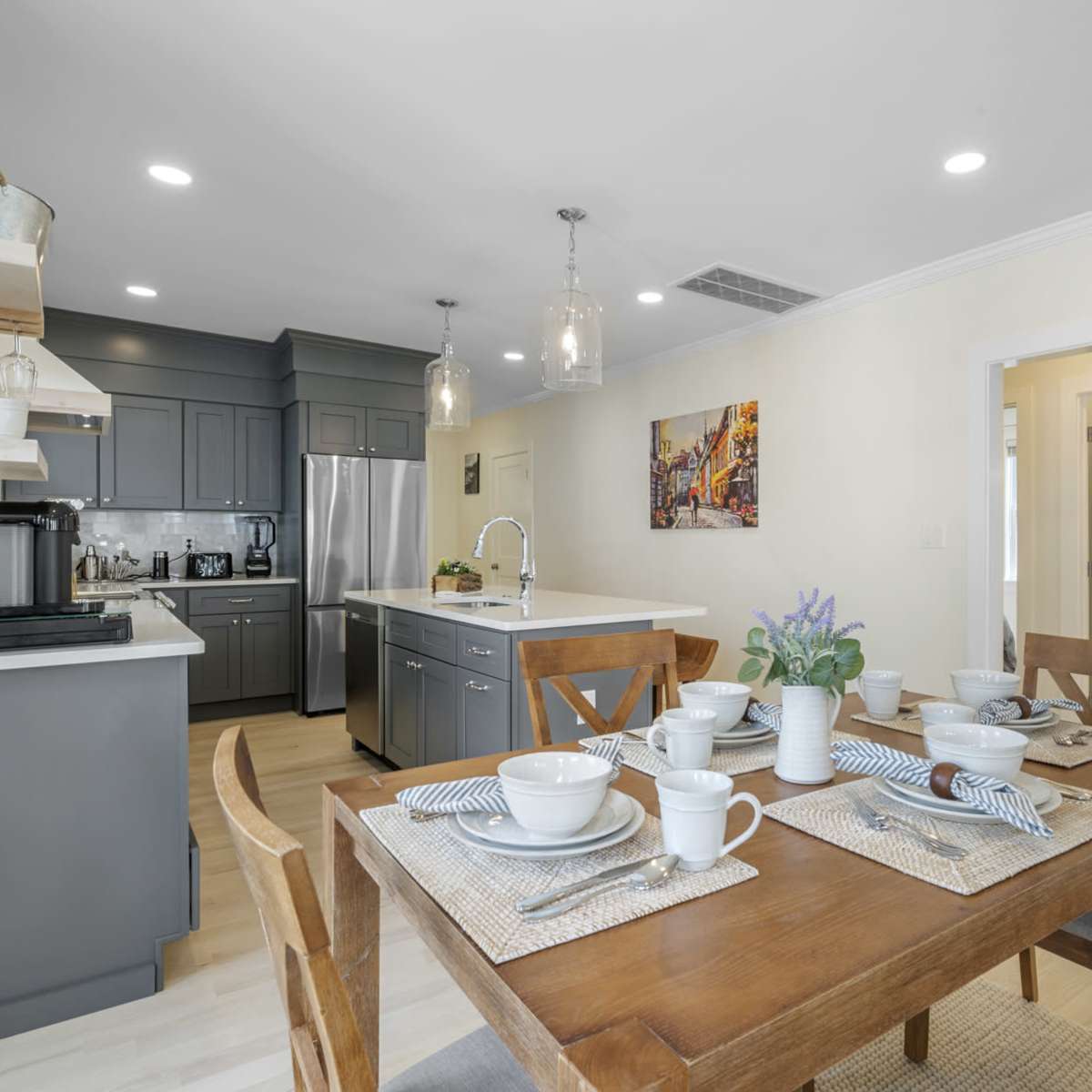 A dining area with a wooden table set for four, adjacent to a modern kitchen featuring gray cabinets and stainless steel appliances.