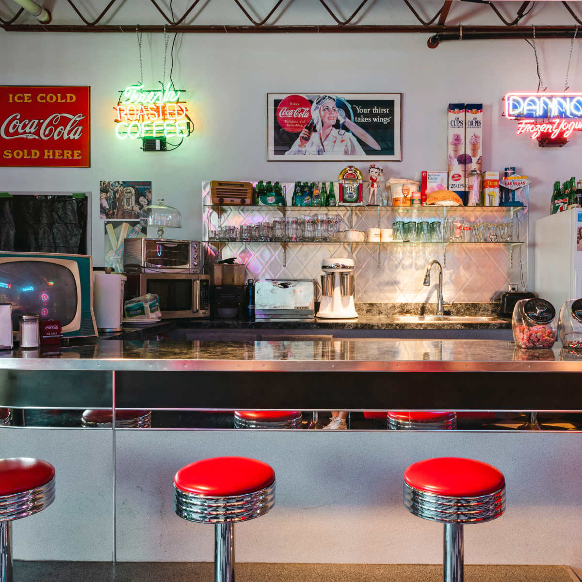 A retro diner interior featuring a chrome counter, red stools, and neon signs advertising Coca-Cola and coffee.