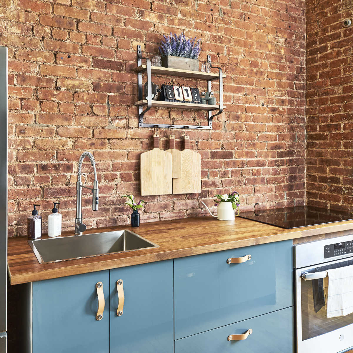 The kitchen features a stainless steel refrigerator, a wooden countertop, a stone sink, and blue cabinetry against a brick wall.