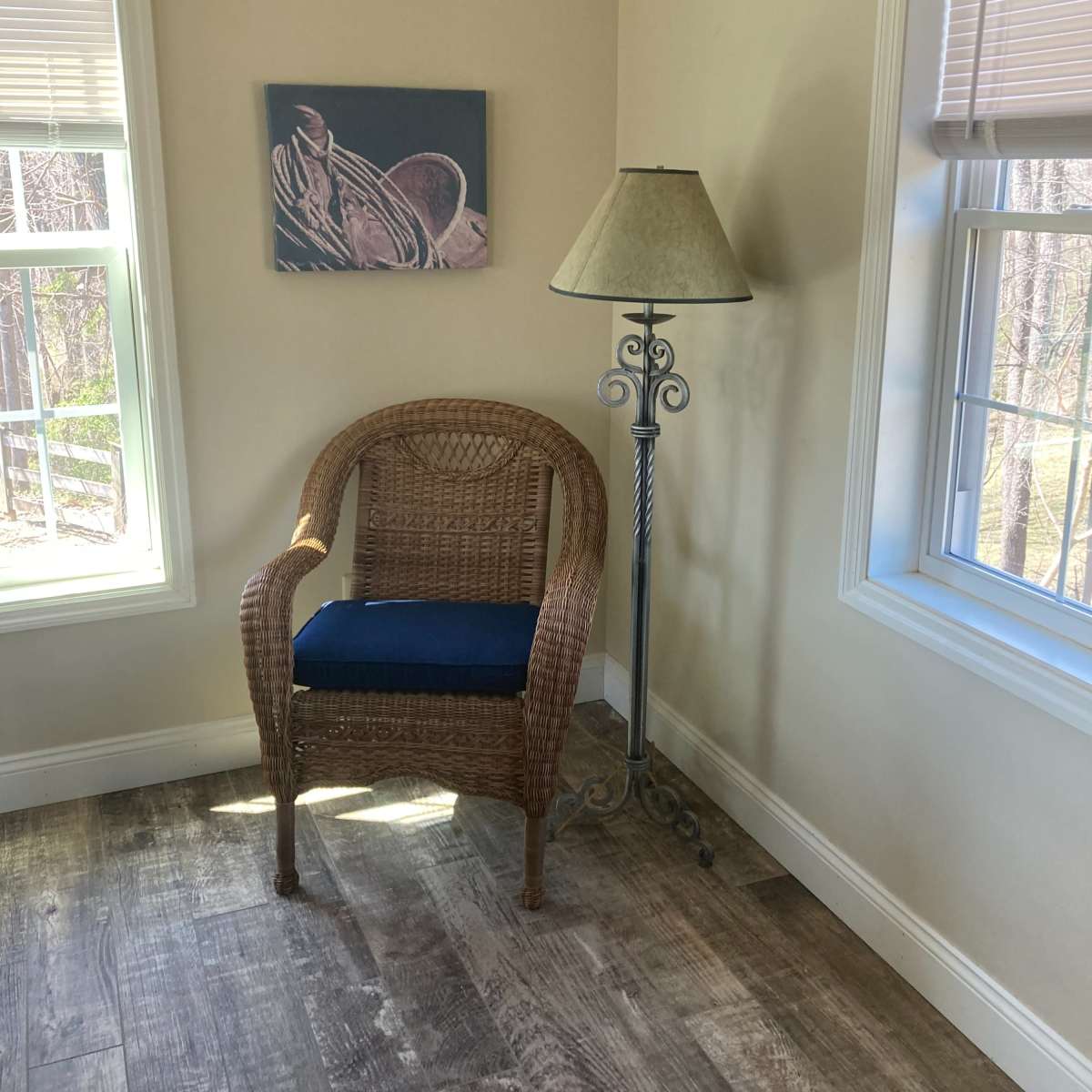 A wicker chair with a blue cushion is positioned next to a floor lamp in a well-lit corner of a room featuring a nature-themed painting on the wall.