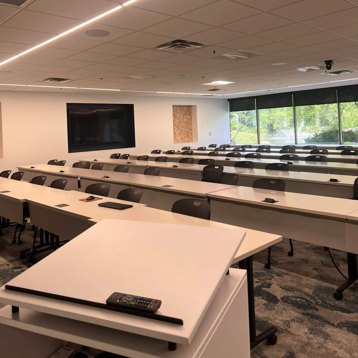 A modern classroom with rows of white tables and chairs, large windows letting in natural light, and a screen mounted on the wall.
