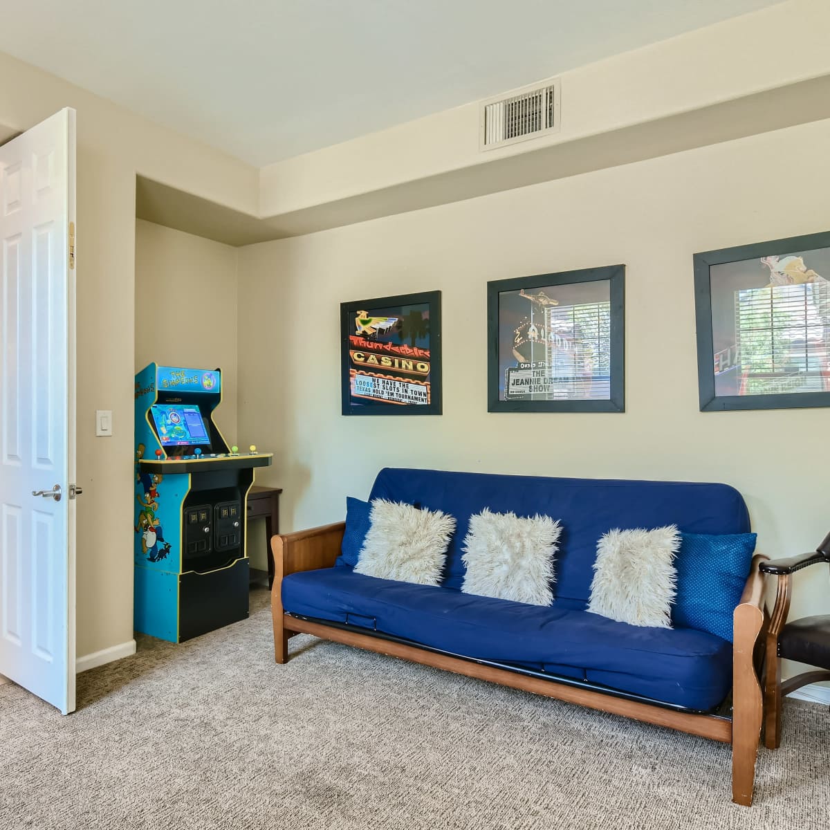 A cozy living room with a blue futon, a black chair, framed art on the walls, and a vintage arcade game machine in the corner.