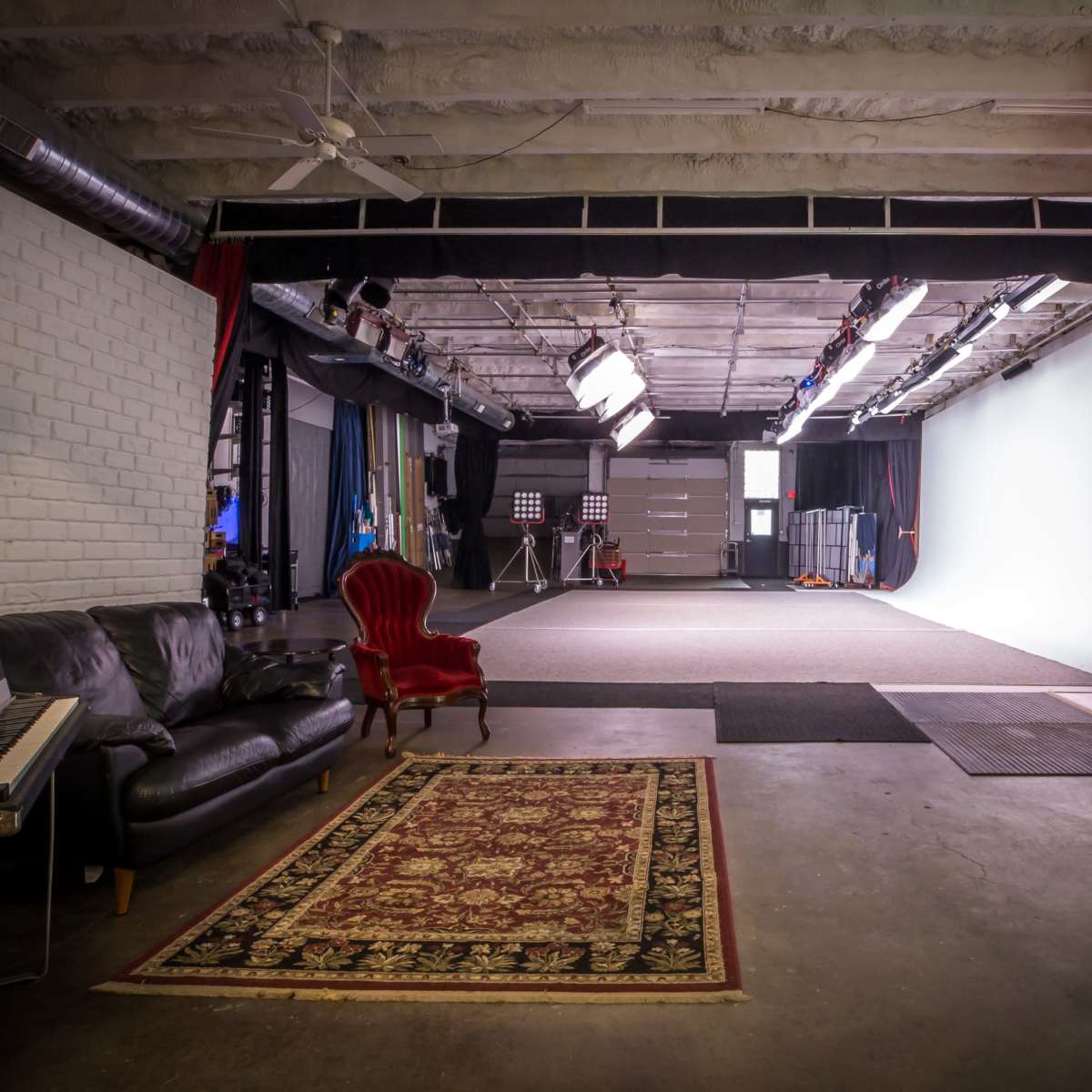 A film studio with a seating area featuring a black sofa and a red chair, leading to a large empty space with a white backdrop and studio lighting.