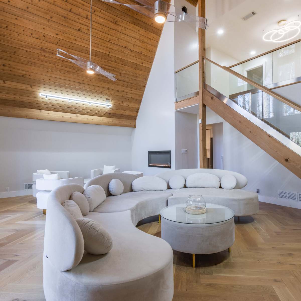 A modern living room with a curved sofa, a glass coffee table, and a staircase leading to an upper level, all accented by wood paneling and large windows.