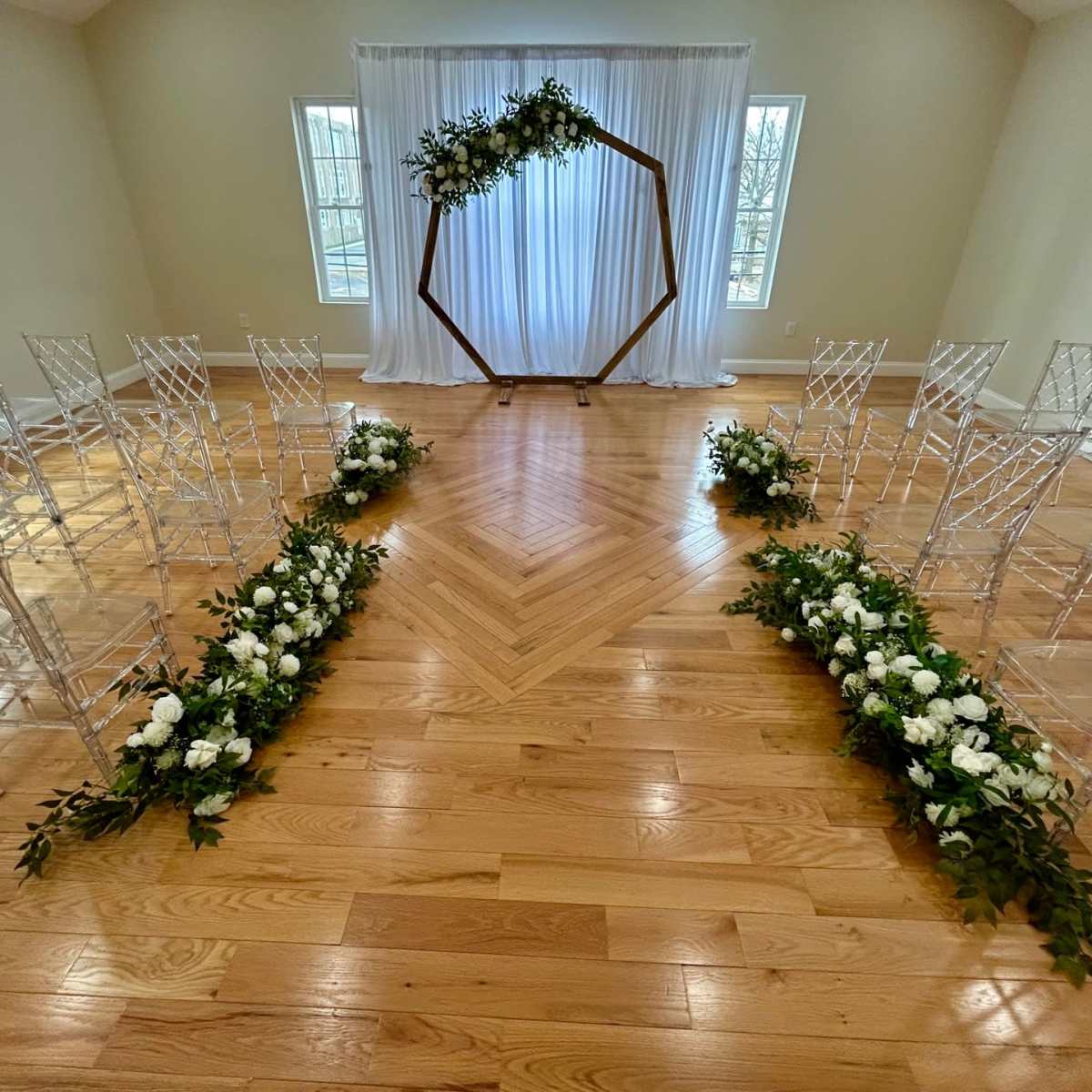 A wedding ceremony setup with transparent chairs arranged in two rows facing an ornate wooden arch decorated with greenery and white flowers, in front of a backdrop of flowing white fabric.