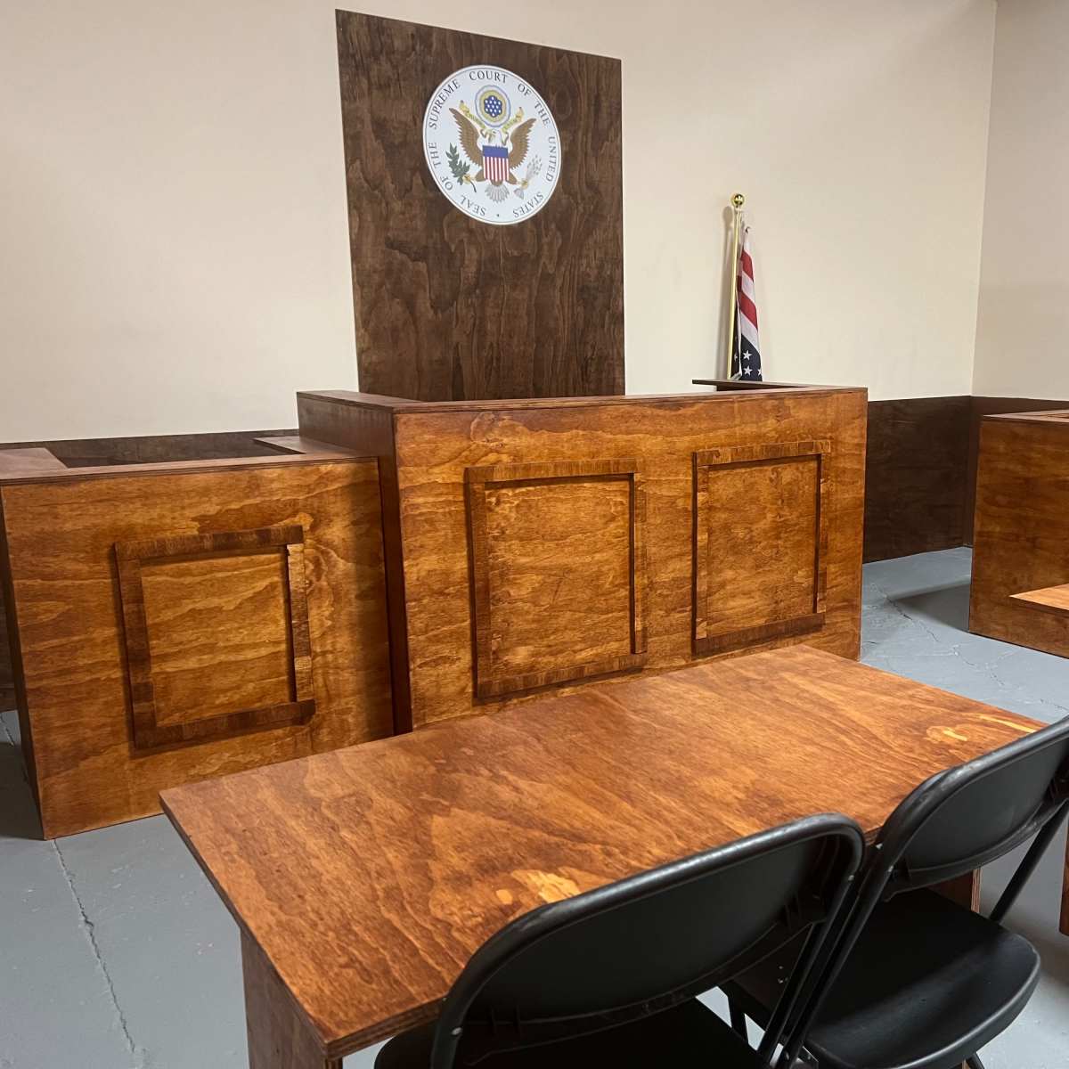 A courtroom setting with wooden furniture, including two witness stands and a table, alongside a United States flag and a seal on the wall.