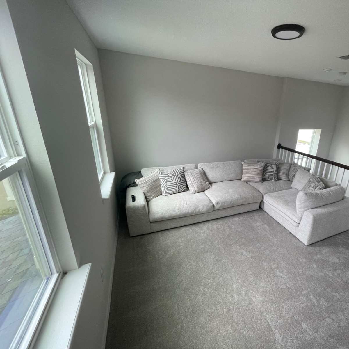 A light gray sectional sofa with decorative pillows is positioned in a well-lit room featuring two windows and a staircase railing.