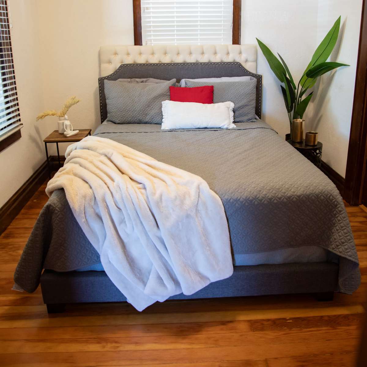 A neatly made bed with a gray bedspread and white decorative pillows in a well-lit bedroom featuring wooden accents and a potted plant.