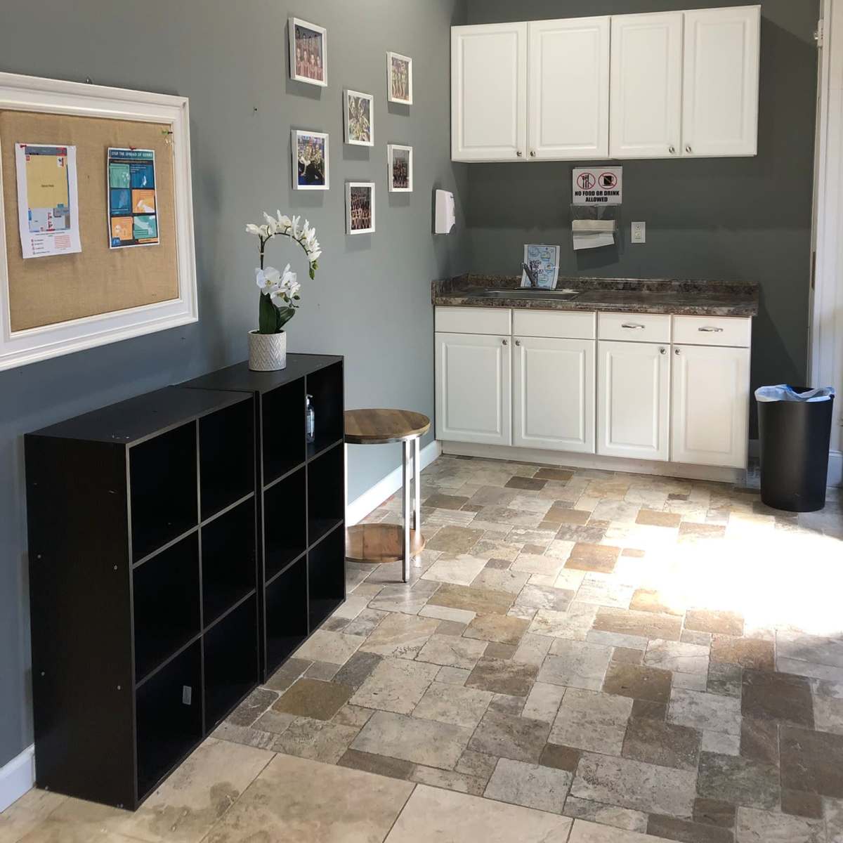 A small interior space with a kitchenette, a bulletin board, framed photos on the wall, and furniture arranged to the side.