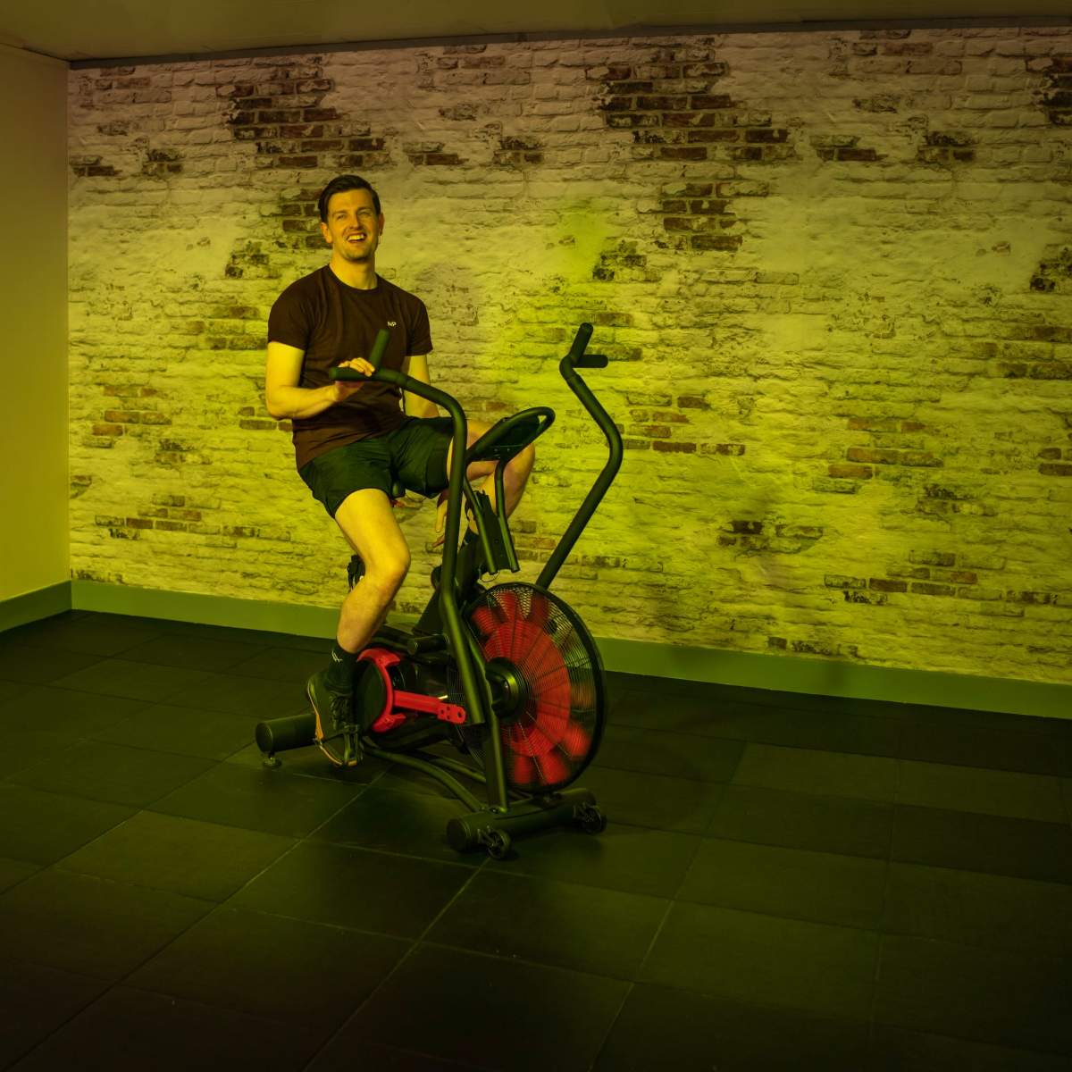 A person exercises on an elliptical machine in a room with a brick wall and a camera set up nearby.