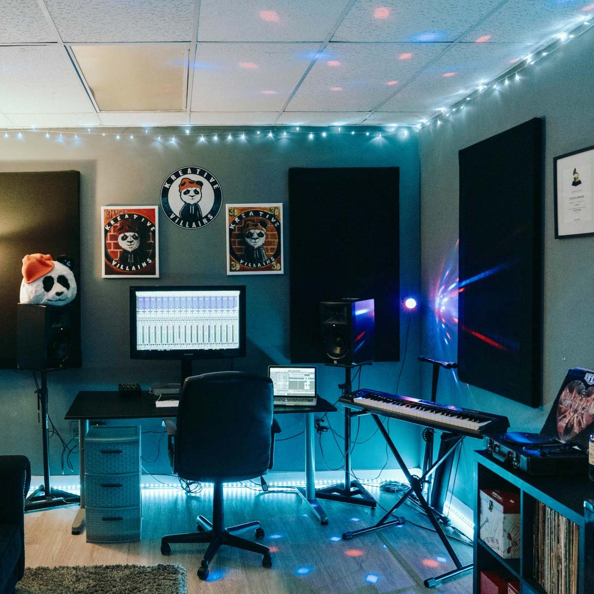 A music studio with a desk set up for production, featuring a computer, speakers, and a keyboard, illuminated by string lights and surrounded by soundproofing panels.