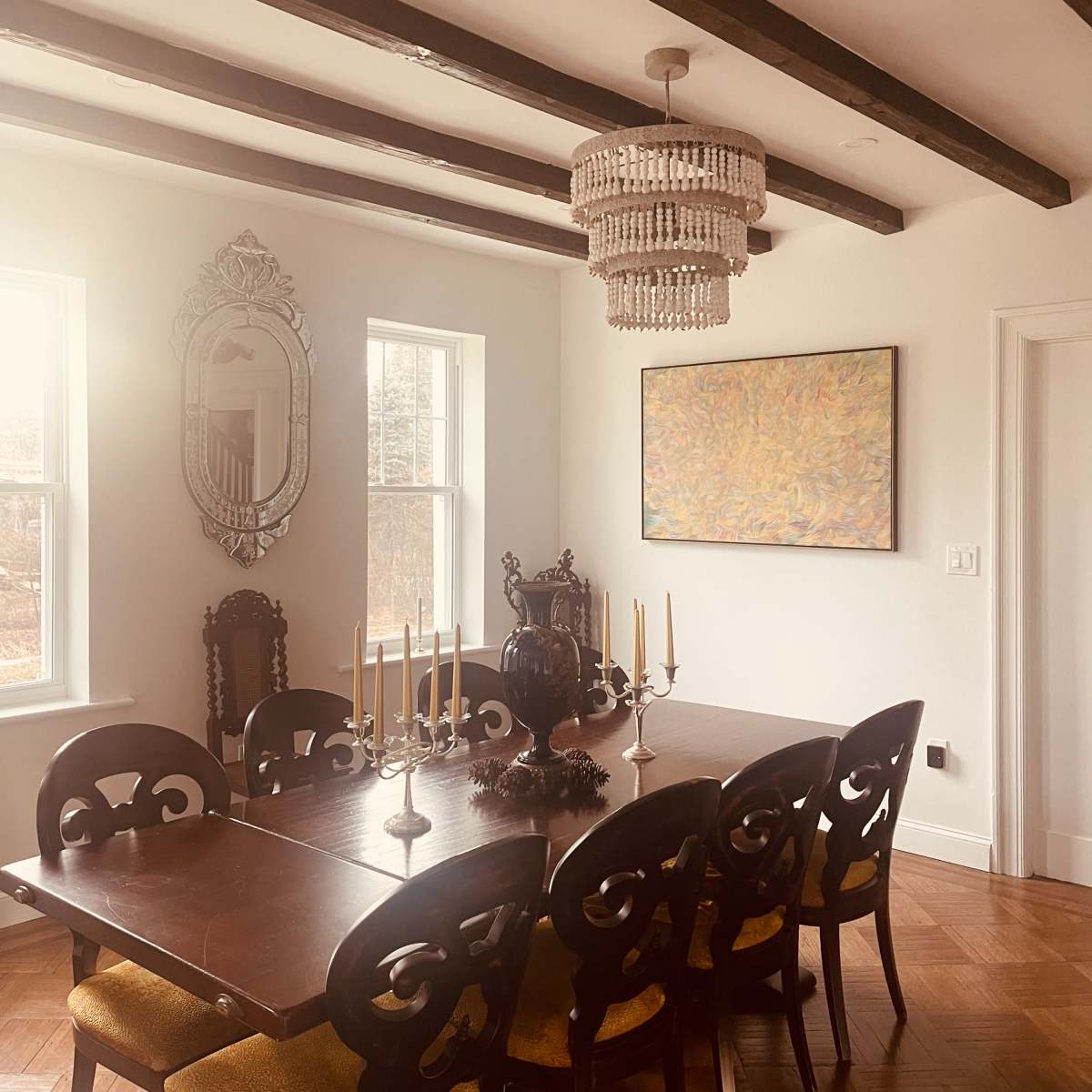 A dining room features a wooden table set for dinner, with a chandelier above and a colorful painting on the wall.