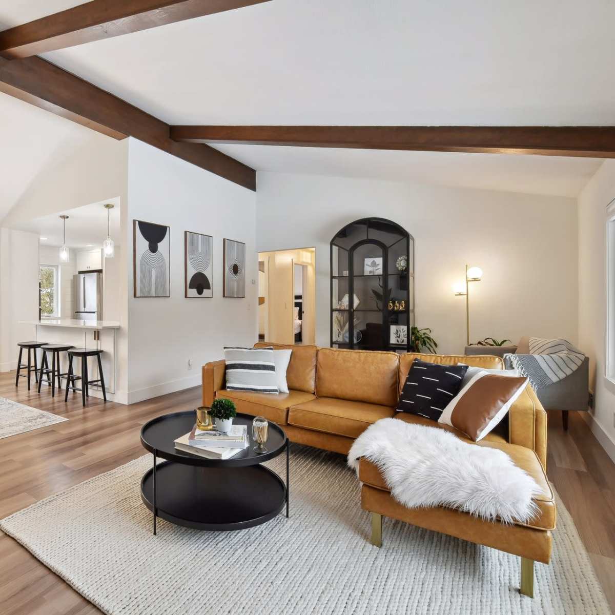 A bright, modern living room with a tan sofa, round coffee table, and wooden beams, connected to a dining area with a black table and chairs.