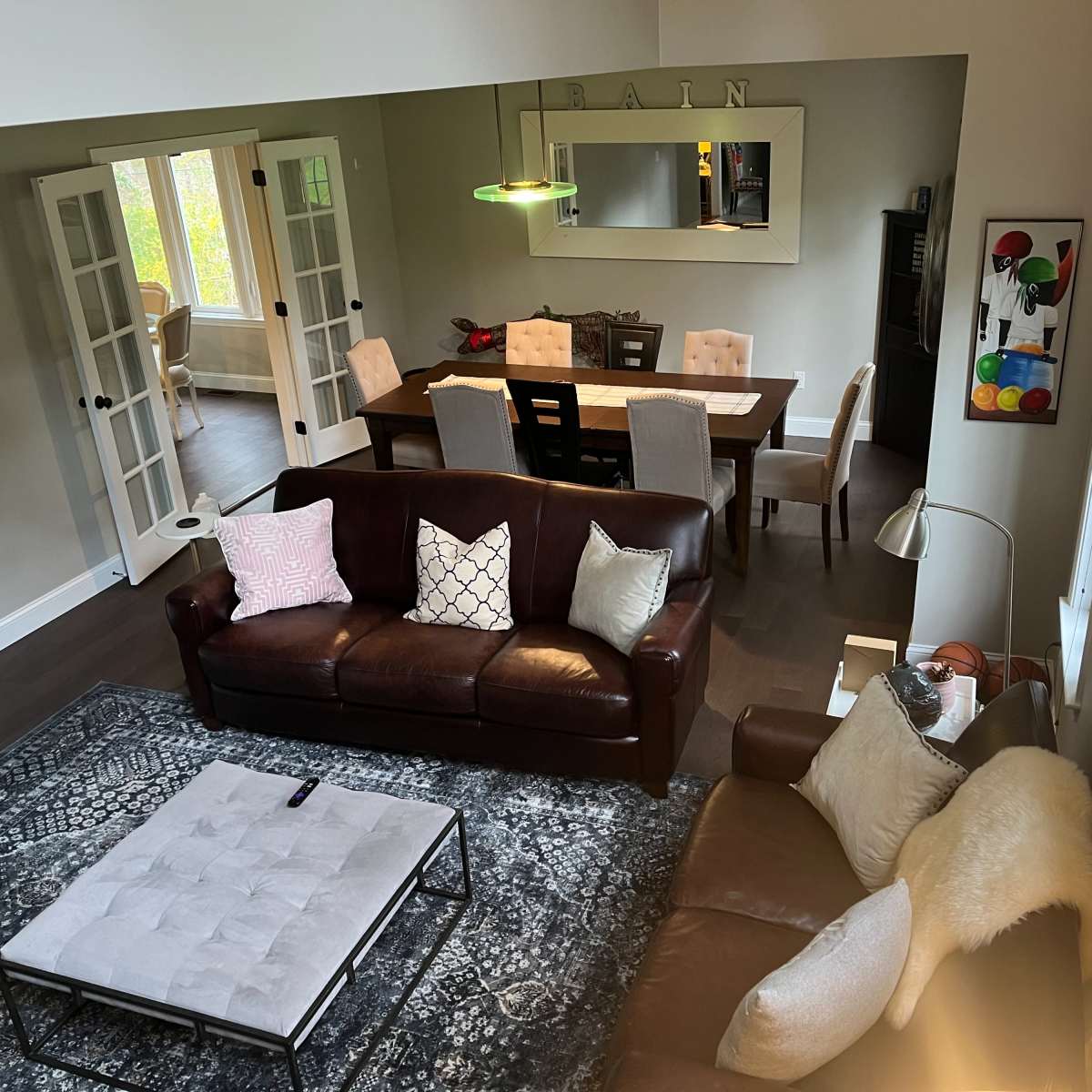 A spacious living room features a brown leather sofa, a light-colored rug, and a dining area with a wooden table set for six.