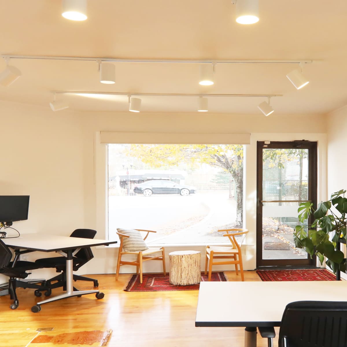 A modern office space featuring two workstations, a small round table with chairs, and large windows allowing natural light.
