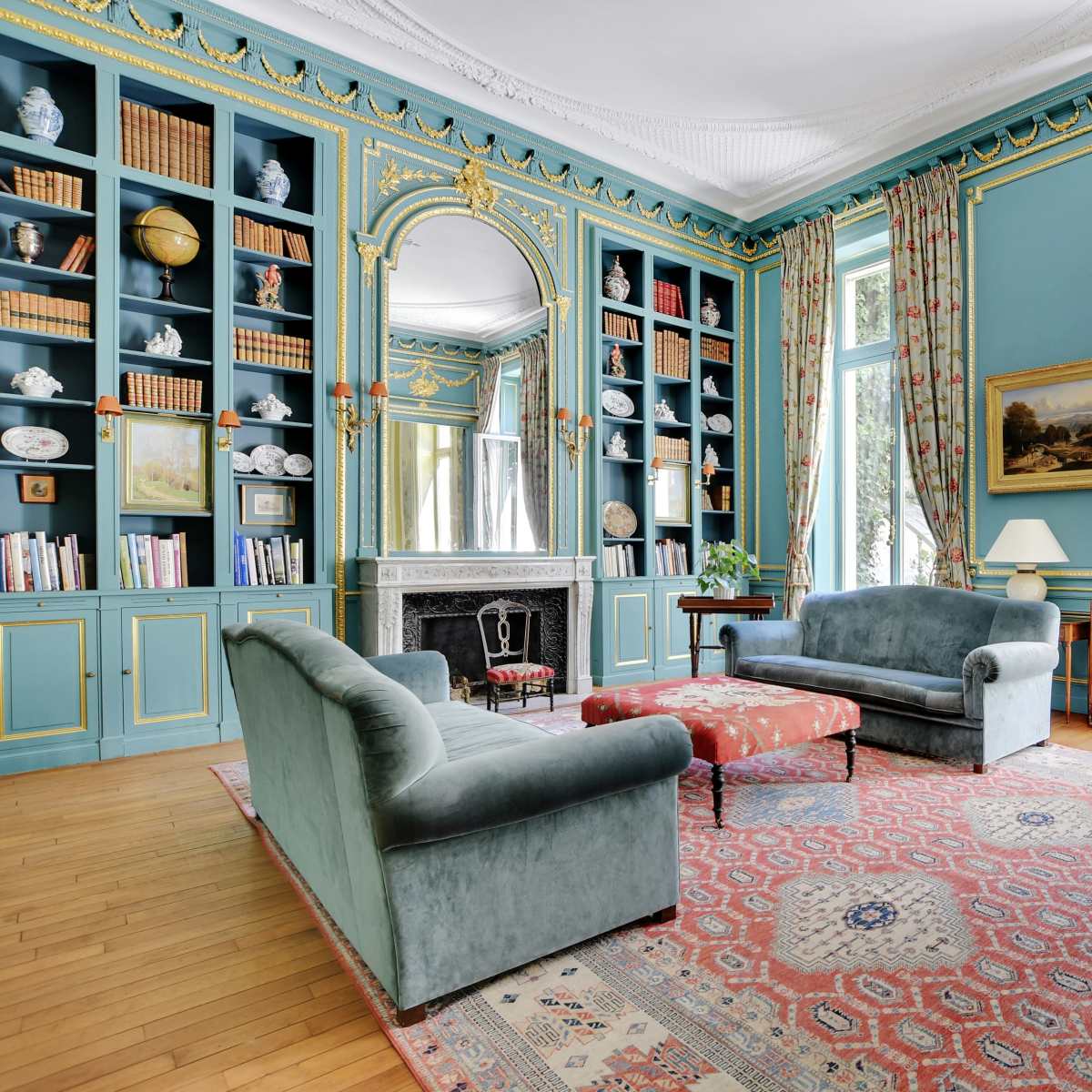 A beautifully furnished living room with light blue walls, large windows, and a central Persian rug.