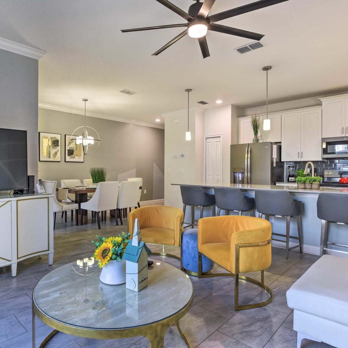 A modern living room with a flat-screen TV, yellow chairs, a coffee table, and a connected dining area featuring a kitchen countertop with bar stools.