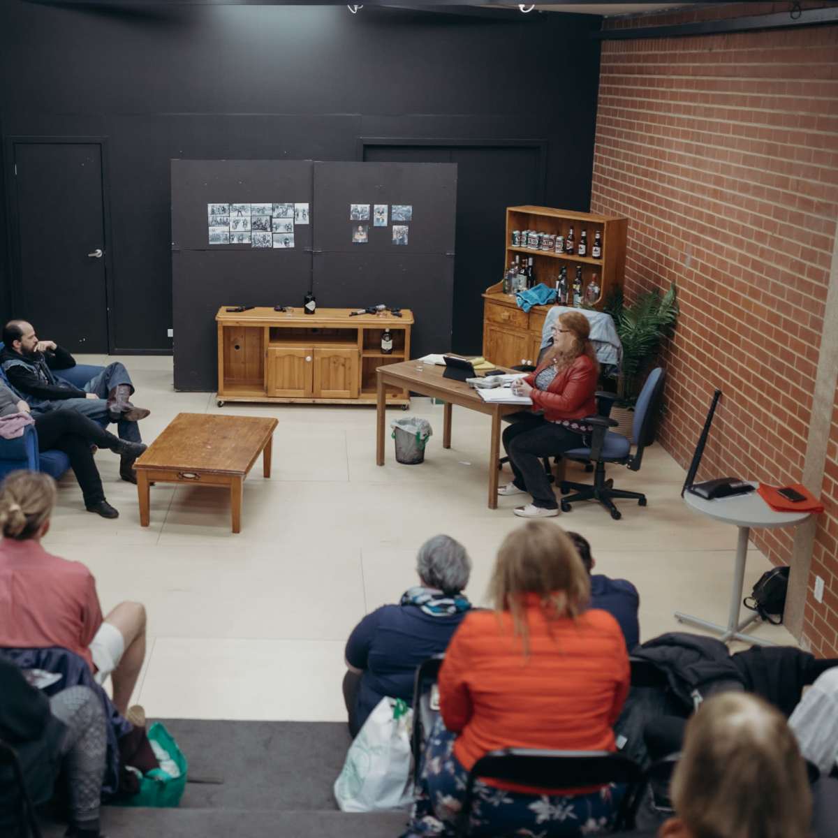 A group of people is seated in a room, observing a discussion taking place between a person at a desk and two individuals on a couch.
