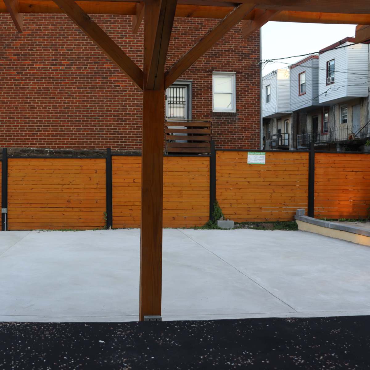 A concrete area with a wooden support structure, bordered by wooden fences and a brick building in the background.