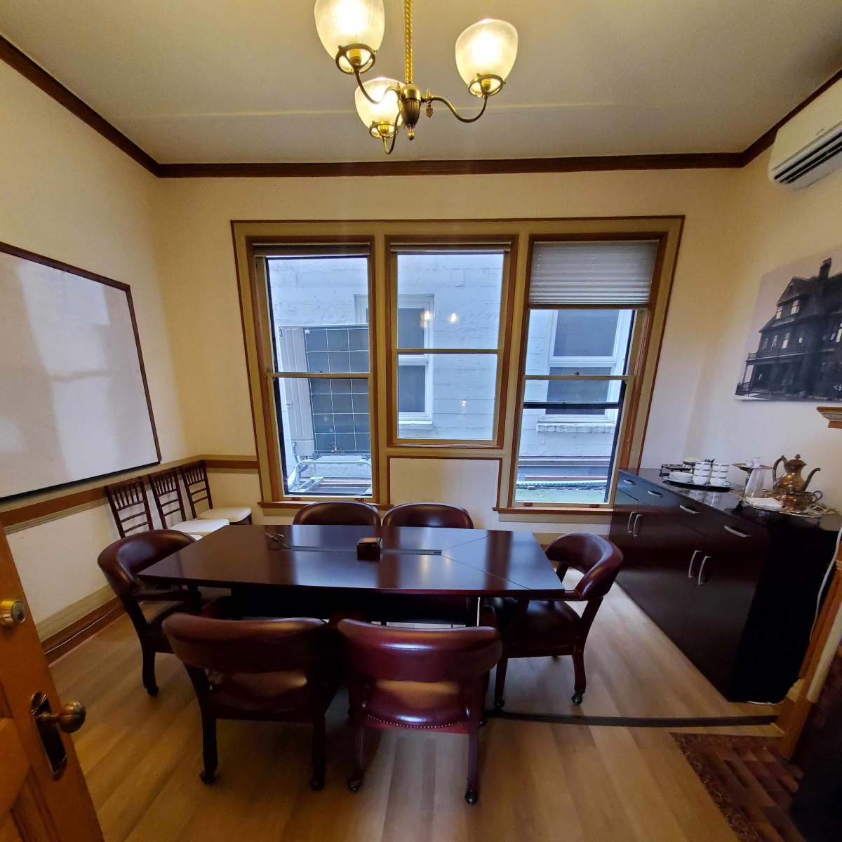A small conference room with a rectangular table surrounded by six wooden chairs, a sideboard, and large windows.